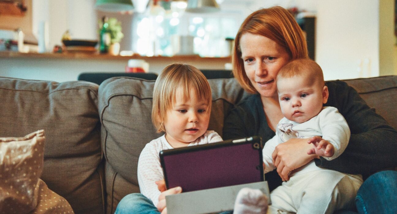 two babies and woman sitting on sofa while holding baby and watching on tablet